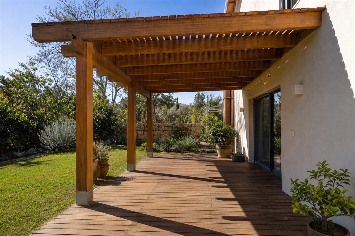 פרגולת עץ מחוברת לקיר הבית, מעל דק בחצר, עם צללים של קורות ההצללה